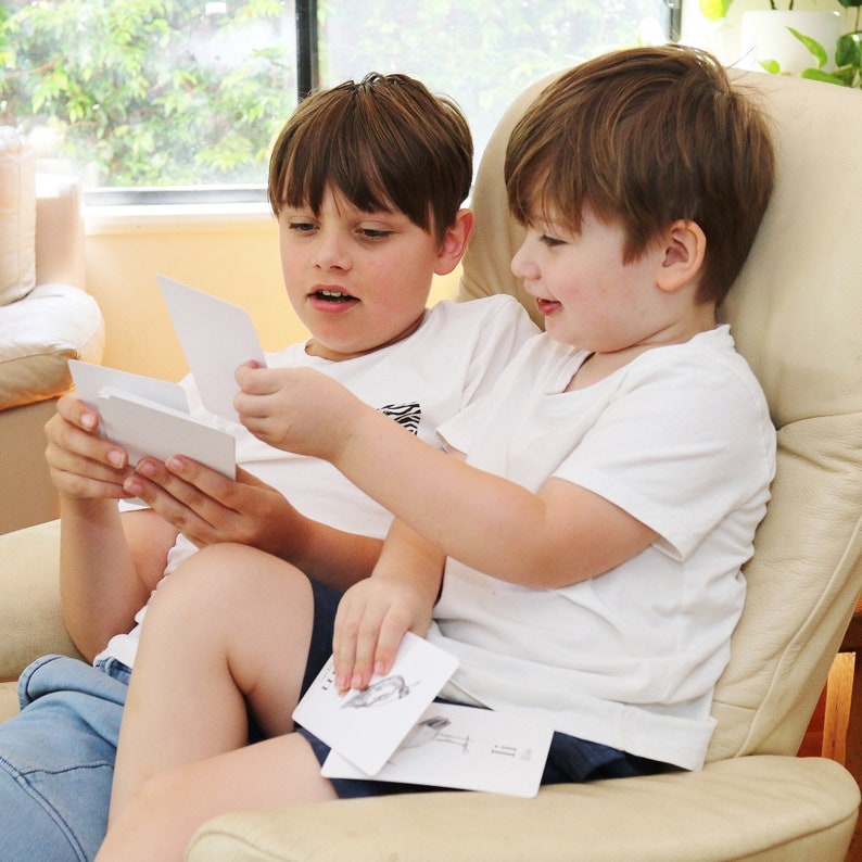 Puede incluir: Dos ni&ntilde;os peque&ntilde;os sentados en un sill&oacute;n beige, mirando cartas con ilustraciones en blanco y negro. El ni&ntilde;o de la izquierda lleva una camiseta blanca con un patr&oacute;n blanco y negro y pantalones vaqueros azules. El ni&ntilde;o de la derecha lleva una camiseta blanca y pantalones vaqueros azules.