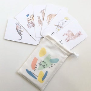 Puede incluir: Un juego de flashcards con animales australianos, incluyendo un canguro, un kookaburra, un wombat y un equidna. Las tarjetas son blancas con ilustraciones coloridas y se empaquetan en una bolsa de cord&oacute;n blanco con el texto "Adventure Awaits".