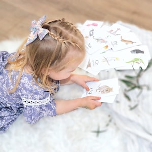 Puede incluir: Una ni&ntilde;a con un vestido floral est&aacute; aprendiendo el alfabeto con flashcards que muestran animales australianos. Las tarjetas muestran ilustraciones de un koala, un kookaburra, un quoll y un casuario.