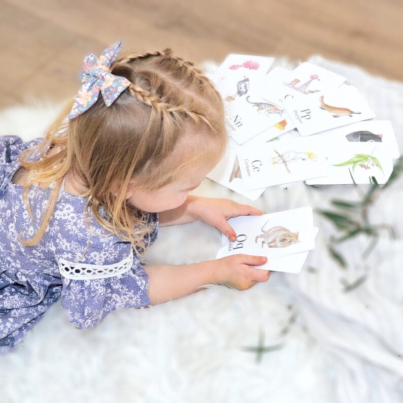 Puede incluir: Una ni&ntilde;a con un vestido floral est&aacute; aprendiendo el alfabeto con flashcards que muestran animales australianos. Las tarjetas muestran ilustraciones de un koala, un kookaburra, un quoll y un casuario.
