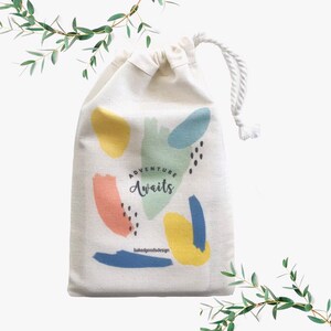 Puede incluir: Una bolsa de tela blanca con cord&oacute;n de ajuste con un dise&ntilde;o abstracto colorido y el texto "Adventure Awaits" impreso en ella. La bolsa est&aacute; decorada con formas abstractas amarillas, azules, verdes y rosas.