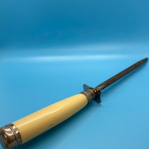 May include: A vintage knife sharpener with a cream-colored handle and a steel sharpening rod. The handle is capped with silver-colored metal. The sharpener is designed for kitchen use and is set against a blue background.