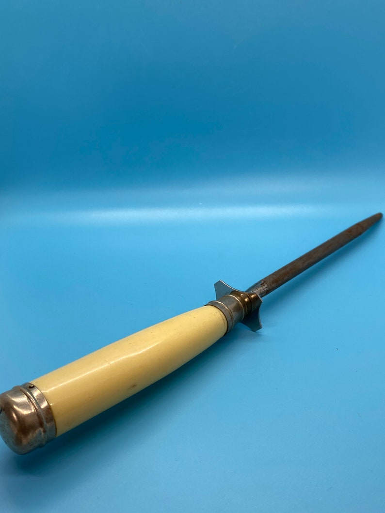 May include: A vintage knife sharpener with a cream-colored handle and a steel sharpening rod. The handle is capped with silver-colored metal. The sharpener is designed for kitchen use and is set against a blue background.