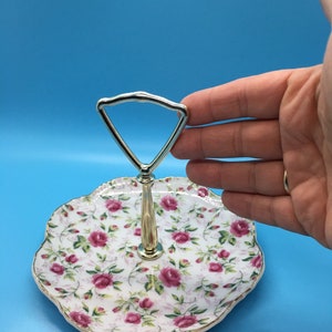 以下が含まれることがあります： バラの花柄の2段サービングトレイ。トレイはスカラップエッジと金色のトリムが付いています。ハンドルはシルバー色です。トレイはおそらく磁器またはセラミック製です。
