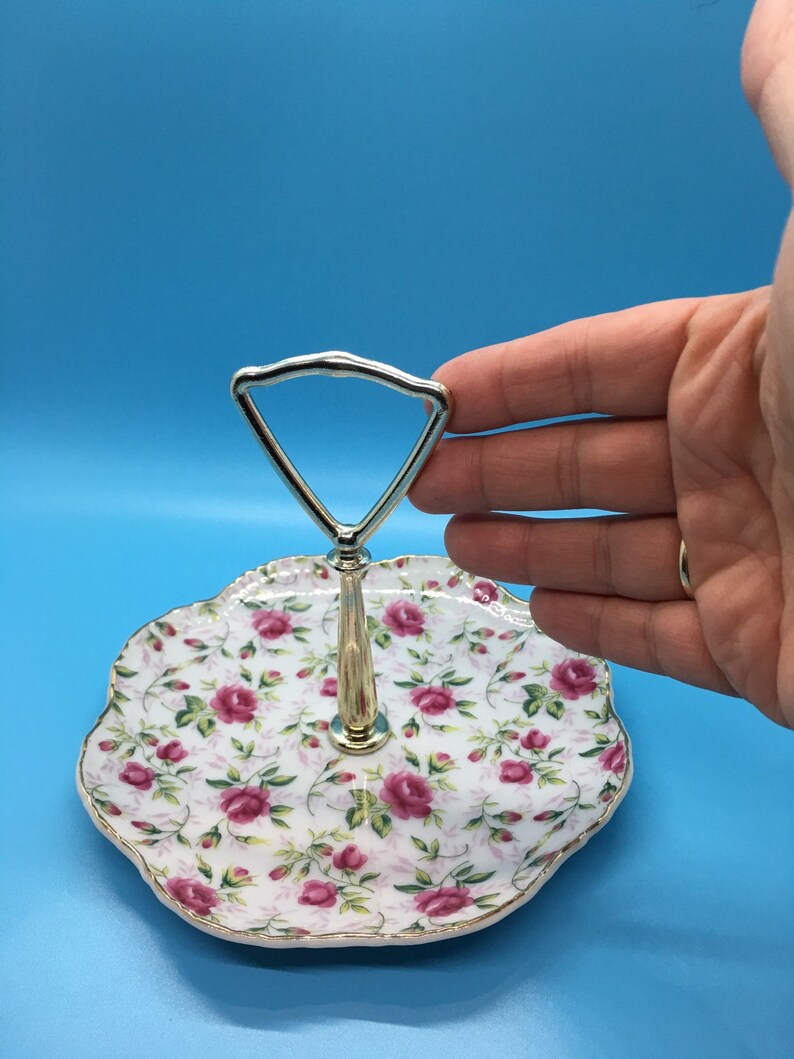 以下が含まれることがあります： バラの花柄の2段サービングトレイ。トレイはスカラップエッジと金色のトリムが付いています。ハンドルはシルバー色です。トレイはおそらく磁器またはセラミック製です。
