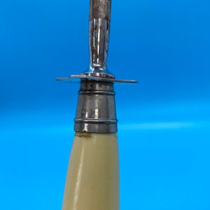 May include: Close-up of a knife sharpener with a cream-colored handle and a metal blade. The blade is attached to the handle with a metal guard. The sharpener is against a blue background.