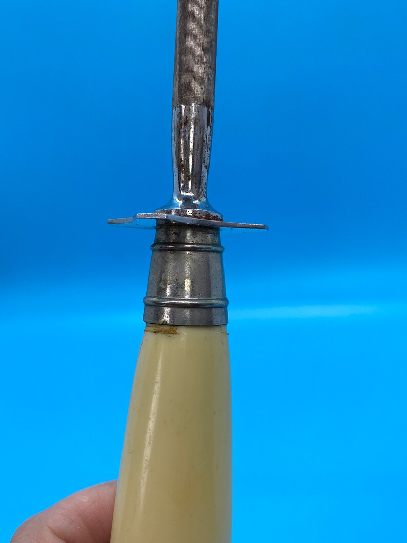 May include: Close-up of a knife sharpener with a cream-colored handle and a metal blade. The blade is attached to the handle with a metal guard. The sharpener is against a blue background.