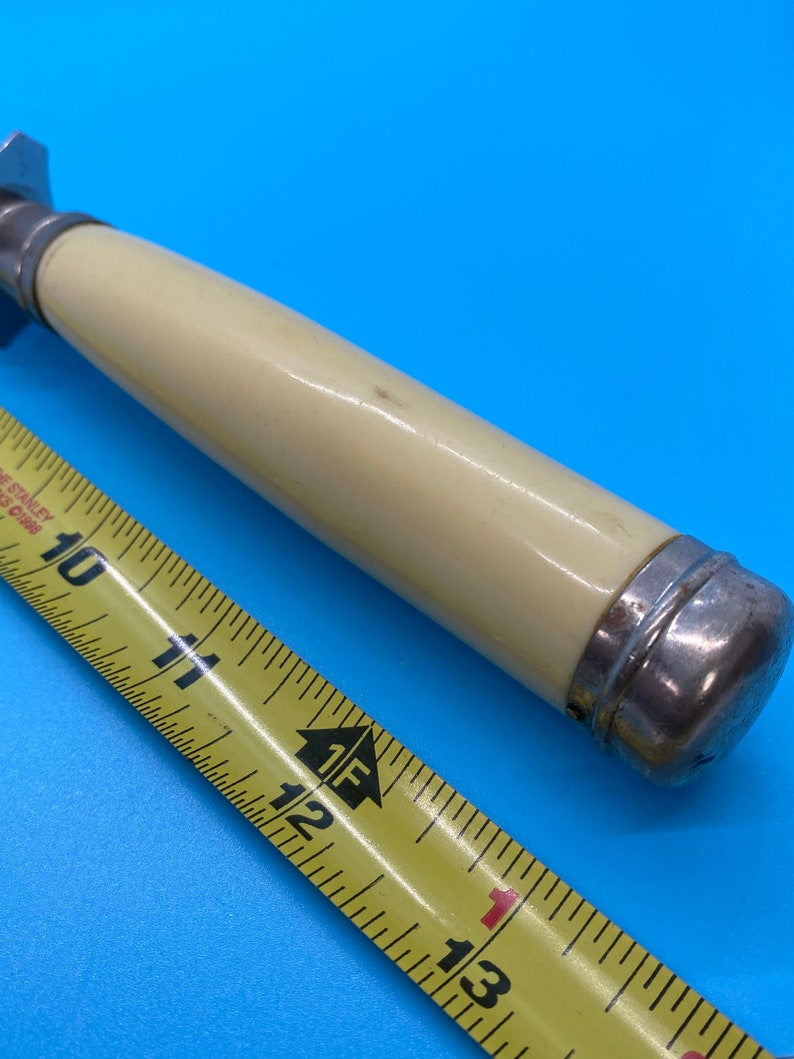 May include: A vintage-style bottle opener with a cream-colored handle and silver-toned metal accents. The handle is approximately 4 inches long. A yellow measuring tape is in the foreground, showing measurements in inches.