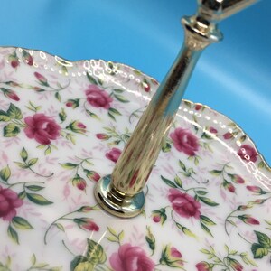 以下が含まれることがあります： バラの花柄の2段サービングトレイ。白い磁器のプレートには、ピンクのバラと緑の葉が飾られています。縁はスカラップで金色のトリムが施されています。金色の金属製のハンドルが層をつないでいます。