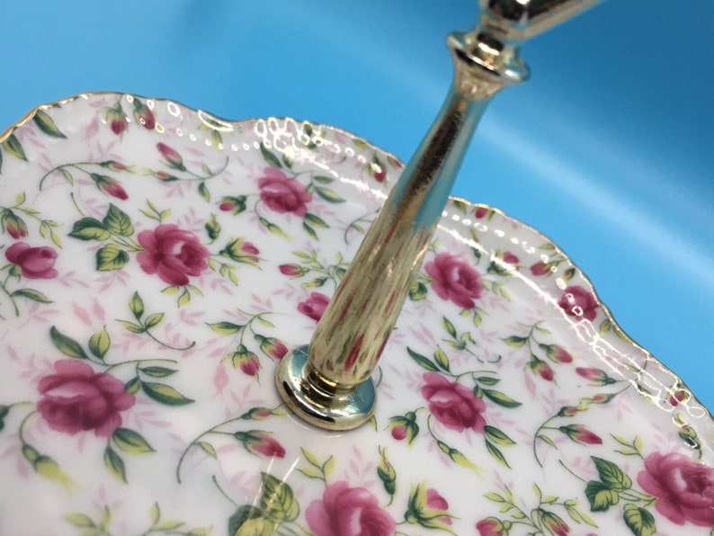 以下が含まれることがあります： バラの花柄の2段サービングトレイ。白い磁器のプレートには、ピンクのバラと緑の葉が飾られています。縁はスカラップで金色のトリムが施されています。金色の金属製のハンドルが層をつないでいます。