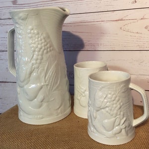 May include: A white ceramic pitcher and two mugs with embossed fruit designs. The pitcher has a handle and a wide mouth. The mugs have handles and are slightly smaller than the pitcher.