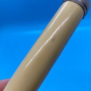May include: A cylindrical object with a cream-colored body and a silver-colored metal cap. The object is angled against a bright blue background. The surface of the object has a slight sheen.
