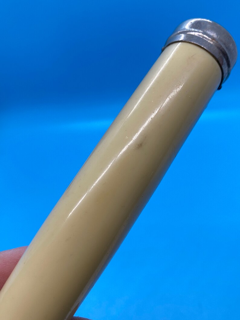 May include: A cylindrical object with a cream-colored body and a silver-colored metal cap. The object is angled against a bright blue background. The surface of the object has a slight sheen.