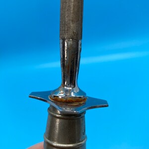 May include: Close-up of a metal knife sharpener with a textured, cylindrical sharpening surface. The handle is off-white with a metal bolster and a star-shaped guard. The tool is designed for sharpening knives.