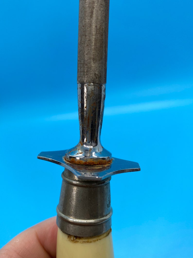 May include: Close-up of a metal knife sharpener with a textured, cylindrical sharpening surface. The handle is off-white with a metal bolster and a star-shaped guard. The tool is designed for sharpening knives.