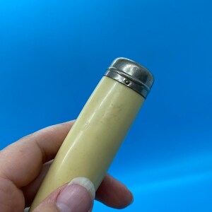 May include: A vintage, cylindrical object with a cream-colored body and a silver-toned metal cap. The object is held against a bright blue background, showcasing its smooth, aged appearance. The cap has a simple, rounded design.