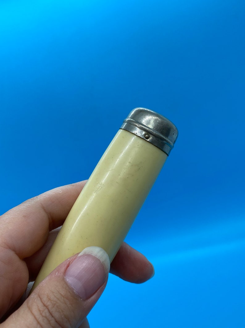 May include: A vintage, cylindrical object with a cream-colored body and a silver-toned metal cap. The object is held against a bright blue background, showcasing its smooth, aged appearance. The cap has a simple, rounded design.