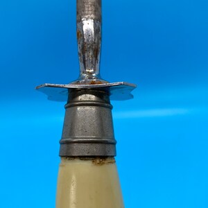 May include: Close-up of a knife sharpener with a cream-colored handle and a metal blade. The blade is attached to the handle with a metal guard. The sharpener is against a blue background.