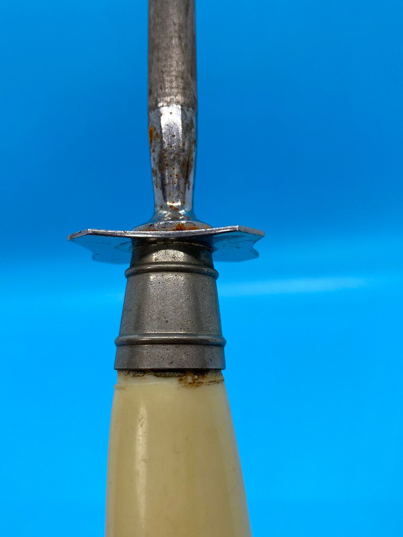 May include: Close-up of a knife sharpener with a cream-colored handle and a metal blade. The blade is attached to the handle with a metal guard. The sharpener is against a blue background.