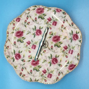 以下が含まれることがあります： バラの花柄の2段サービングトレイ。白い磁器のプレートは、ピンクのバラと緑の葉のデザインで、金のトリムが施されています。2つのプレートはシルバーのハンドルで接続されています。トレイは青色の背景にあります。