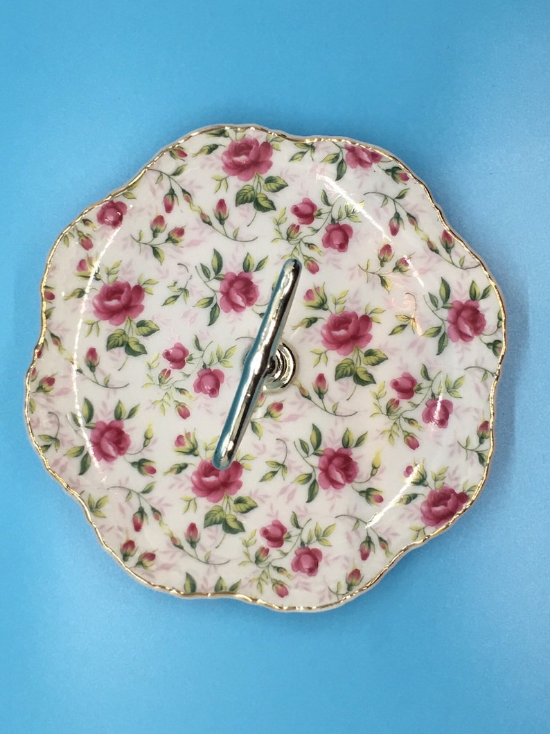 以下が含まれることがあります： バラの花柄の2段サービングトレイ。白い磁器のプレートは、ピンクのバラと緑の葉のデザインで、金のトリムが施されています。2つのプレートはシルバーのハンドルで接続されています。トレイは青色の背景にあります。