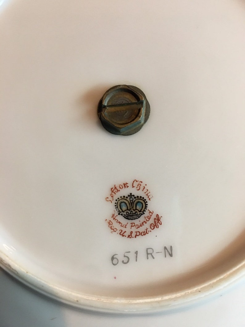 以下が含まれることがあります： 白い磁器の皿のクローズアップ。金属製のネジと、赤と黒で「Seton China Hand Painted Reg. U.S. Pat. Off.」の文字が書かれています。皿には「651 R-N」の数字も印刷されています。