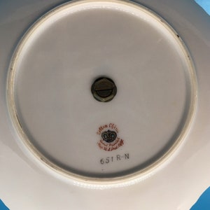 以下が含まれることがあります： 白い波状の縁が付いた磁器のプレート。裏面には、中央に真鍮製の継手と、「Seton China Hand Painted Reg. U.S. Pat. Off.」と番号「651 R-N」と書かれた装飾的なエンブレムが付いています。