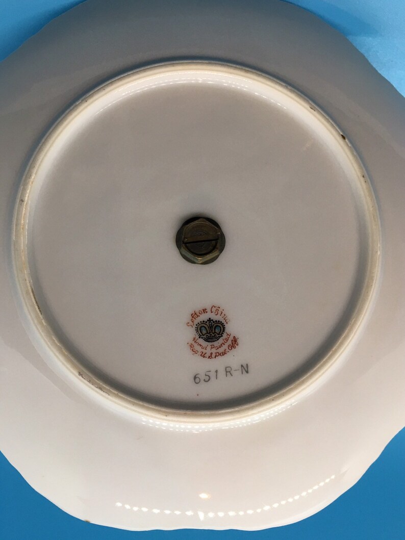 以下が含まれることがあります： 白い波状の縁が付いた磁器のプレート。裏面には、中央に真鍮製の継手と、「Seton China Hand Painted Reg. U.S. Pat. Off.」と番号「651 R-N」と書かれた装飾的なエンブレムが付いています。