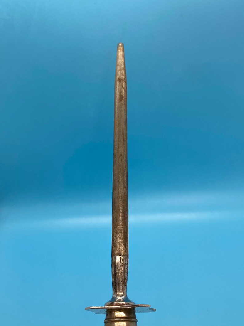 May include: A metal knife sharpener with a long, tapered steel rod. The rod is a dull silver color with signs of wear. The base of the sharpener has a decorative, star-shaped design. The background is a solid light blue.