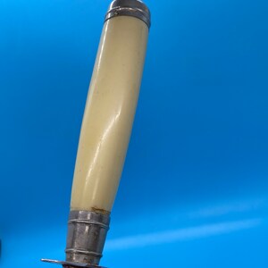 May include: Close-up of a carving knife with a cream-colored handle and a silver metal bolster and blade. The handle is cylindrical and slightly curved, with a silver cap at the top. The blade has a small guard.