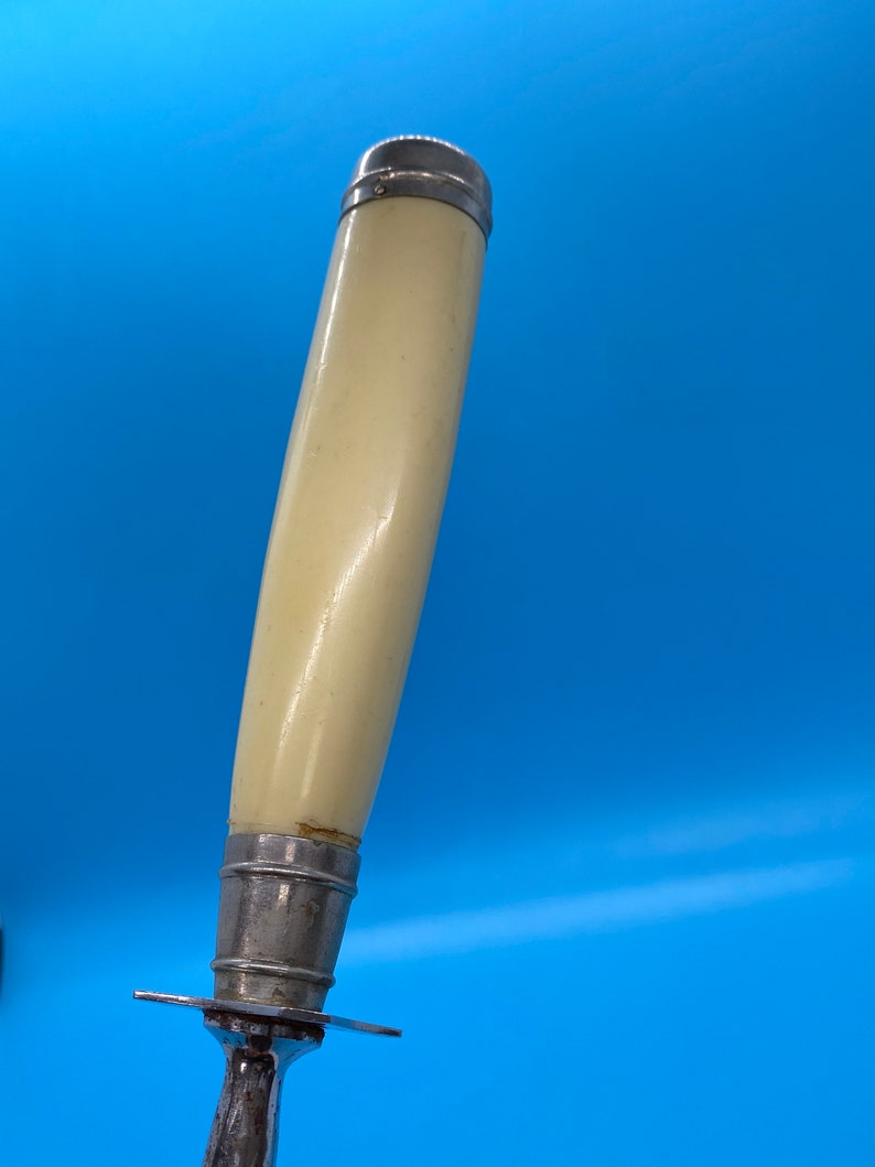 May include: Close-up of a carving knife with a cream-colored handle and a silver metal bolster and blade. The handle is cylindrical and slightly curved, with a silver cap at the top. The blade has a small guard.