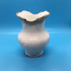May include: A white ceramic vase with a ruffled rim and a gold trim. The vase has a bulbous body and a pedestal base. The background is a solid light blue.