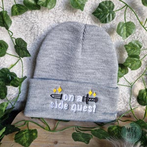 Op de afbeelding: Grijze gebreide beanie met een zwart geborduurd zwaard en de tekst "on a side quest" in wit.