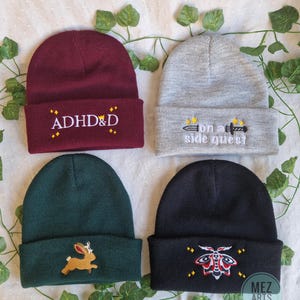 Op de afbeelding: Vier mutsen in bordeaux, grijs, groen en zwart. De bordeauxrode muts heeft de witte borduring "ADHD&D". De grijze muts heeft de witte borduring "on a side quest". De groene muts heeft een bruin konijnenpatch. De zwarte muts heeft een motief.