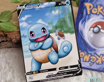 May include: A Squirtle V Pokémon trading card with a blue and tan colour scheme. The card features a cartoon image of Squirtle on a beach. The card has the text "Squirtle V" and "HP 70". Another Pokémon card is visible in the background.