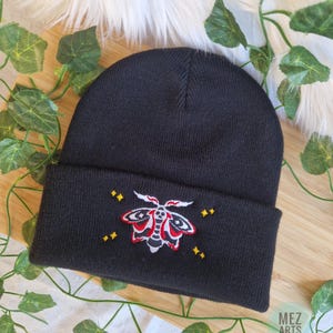 Op de afbeelding: Zwarte beanie met een omgeslagen rand, met een geborduurd motontwerp in rood, wit en geel. De beanie is gemaakt van zacht, gebreid materiaal. De tekst "MEZ ARTS" is zichtbaar in de rechter benedenhoek.