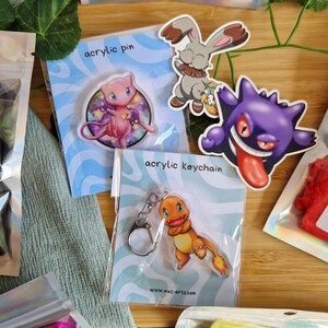 Peut inclure: Diverses épingles et porte-clés en acrylique colorés représentant des personnages de dessins animés. Les épingles incluent une créature rose, un lapin gris et un fantôme violet. Un porte-clés avec un lézard orange est également visible.