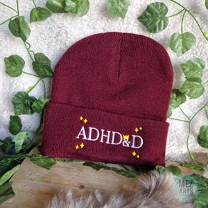 Op de afbeelding: Een bordeauxrode beanie met de tekst "ADHD&D" geborduurd in wit met gele diamantvormen rond de tekst.