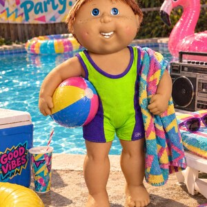 May include: A Cabbage Patch Kids doll with brown hair and blue eyes wearing a lime green and purple swimsuit. The doll holds a multi-colored beach ball and a patterned towel. The doll has freckles and a wide smile with visible teeth.