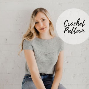 May include: A woman wearing a gray crochet top with short sleeves. The top has a loose fit and a slightly boat neck. The text "Crochet Pattern" is written in a white circle in the upper right corner of the image.
