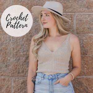 May include: A woman wearing a beige crochet tank top with a v-neck and a white hat. The tank top is cropped and the woman is wearing blue jeans.