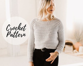 May include: A close-up of a woman wearing a light gray crochet sweater with long sleeves. The sweater has a ribbed texture and a slightly cropped length. The text "Crochet Pattern" is visible in a circle in the foreground.
