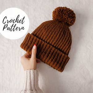 Könnte beinhalten: Eine braune Häkelmütze mit einem flauschigen Bommel oben. Die Mütze wird von einer Hand mit rotem Nagellack gehalten.