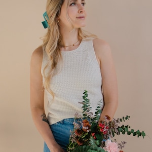 A model holding a floral bouquet and wearing a white crocheted tank top, looking off to the side.