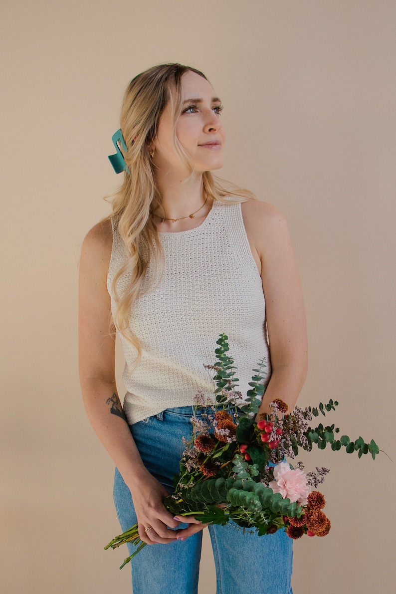 A model holding a floral bouquet and wearing a white crocheted tank top, looking off to the side.