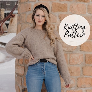 May include: A woman wearing a brown knitted sweater, blue jeans, and a black headband. The sweater has a loose fit and long sleeves. The woman is standing in front of a brick wall with the text "Knitting Pattern" in a circle.