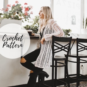 May include: A white crochet cardigan with a lacy pattern. The cardigan is worn by a person sitting on a black chair in a cafe setting.