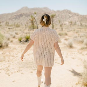 May include: A woman wearing a white crochet dress and white cowboy boots walks through a desert landscape.
