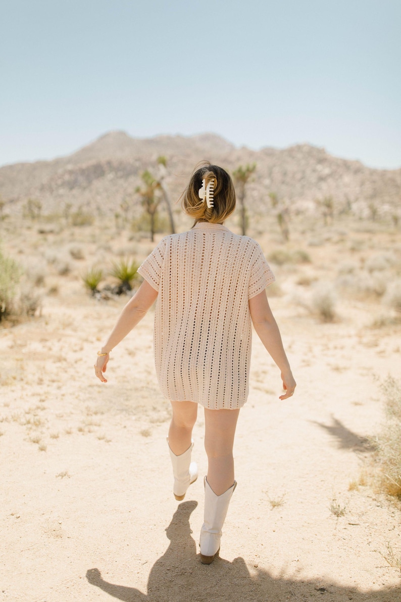 May include: A woman wearing a white crochet dress and white cowboy boots walks through a desert landscape.
