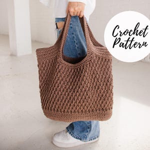 以下が含まれることがあります： 茶色の編み物トートバッグで、質感のあるステッチパターンが施されています。バッグは、青いジーンズを履いた人が持っています。画像の右上隅には、円の中に「Crochet Pattern」という文字が書かれています。